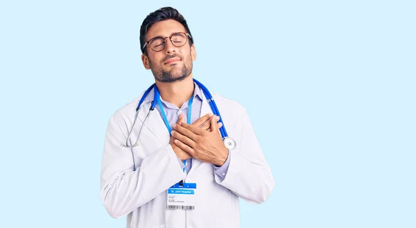 医師の制服を着た若いヒスパニック系の男が目を閉じて胸に手で笑みを浮かべて聴診器と顔に感謝ジェスチャー 健康の概念 — ストック写真