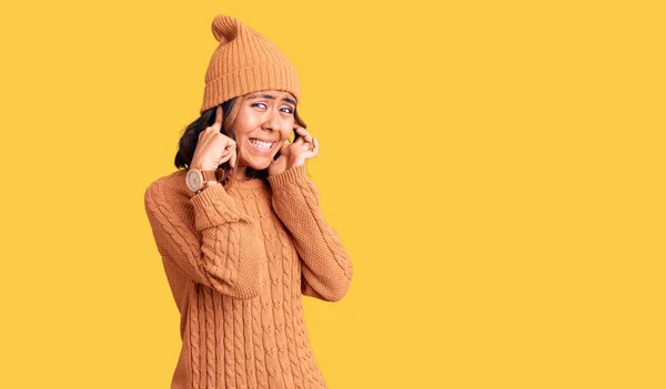 ウールのセーターと冬の帽子を身に着けている若い美しい混合レースの女性は 大きな音楽のノイズのためのいらいらした表情で指で耳を覆う 聴覚障害の概念 — ストック写真