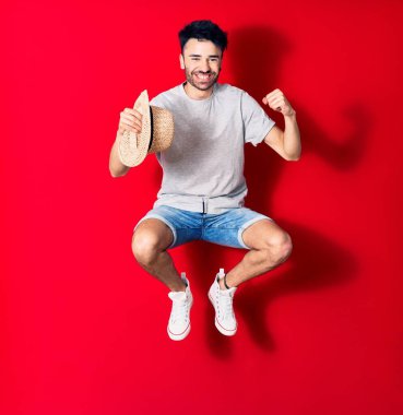 Genç, yakışıklı, sıradan kıyafetler giyen, mutlu gülümseyen bir İspanyol. Yüzünde tebessümle zıplayarak yaz şapkası tutarak izole edilmiş kırmızı arka planda yumruğunu havaya kaldırarak kutluyor..