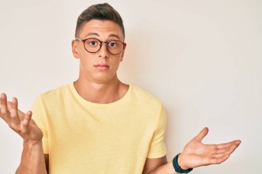 Sıradan kıyafetler giyen, gözlüklü, kafası karışmış, elleri ve kolları havada bir İspanyol çocuk. şüphe kavramı. 