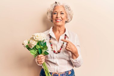 Yaşlı gri saçlı kadın elinde çiçekler tutarken mutlu mesut eli ve parmağı ile işaret ediyor. 
