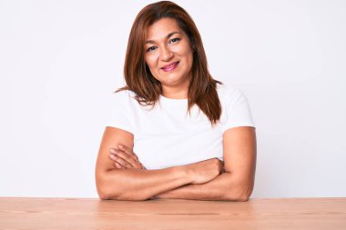 Orta yaşlı esmer, beyaz tişört giyen, masanın üzerinde oturan mutlu yüzlü, kollarını kavuşturup kameraya bakan Latin bir kadın. pozitif kişi. 