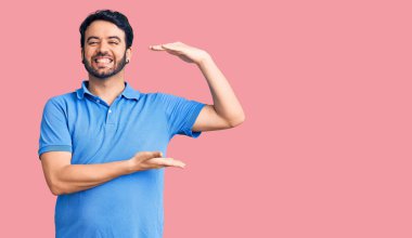 Latin asıllı genç adam, sıradan kıyafetler giyiyor. Elleriyle işaret ediyor. Ölçü sembolü, büyük boy işareti. Kameraya bakıp gülümsüyor. ölçüm kavramı. 