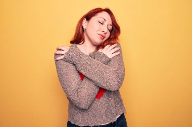 Genç, güzel, kızıl saçlı bir kadın, sarı arka planda gündelik bir kazak giyiyor, kendine sarılıyor, mutlu ve pozitif, kendinden emin gülümsüyor. Sevgi ve özgüven.