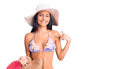 Bikini giyen ve şapka takan genç, güzel bir Latin kız elinde torbayla gülümseyen ve gururlu birini işaret ediyor. 