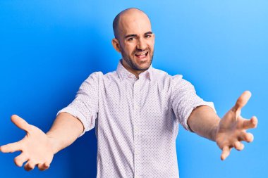 Zarif bir gömlek giymiş genç ve kel bir adam kameraya bakıp sarılmak için kollarını açmış gülümsüyor. Mutluluğu kucaklayan neşeli bir ifade.. 