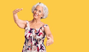 Son sınıftaki gri saçlı kadın, günlük kıyafetler giyip kameraya bakıyor ve sarılmak için kollarını açmış gülümsüyor. Mutluluğu kucaklayan neşeli bir ifade.. 