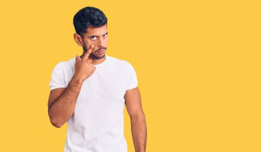 Gündelik kıyafetler giyen genç latin adam senin jestini seyrediyor, şüpheli bir ifade. 