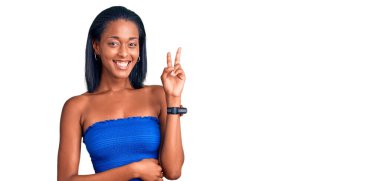 Genç Afro-Amerikan kadın günlük yaz kıyafetleri giyip kameraya gülümseyen ve parmaklarıyla zafer işareti yapan. İki numara.. 