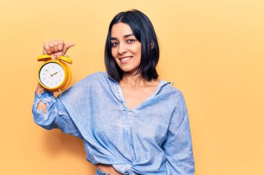 Çalar saati tutan genç ve güzel Latin bir kadın. Pozitif ve mutlu görünüyor. Duruyor ve gülümsüyor. 