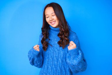 Genç, güzel Çinli bir kız, sıradan bir kazak giyiyor. Çok mutlu ve heyecanlı. Kazanan jestler yapıyor, kolları havada, gülümsüyor ve başarı için bağırıyor. kutlama konsepti. 