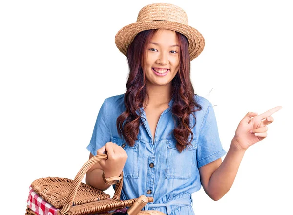 年轻美丽的中国姑娘戴着夏帽 提着野餐柳条篮 手里拿着面包 笑容满面 手指手画脚 — 图库照片