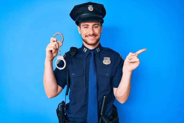 Young caucasian man wearing police uniform holding handcuffs smiling happy pointing with hand and finger to the side