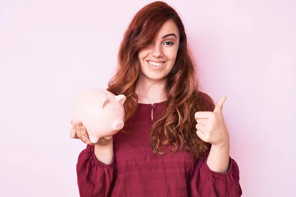 Joven Hermosa Mujer Sosteniendo Alcancía Sonriendo Feliz Positivo Pulgar Hacia —  Fotos de Stock