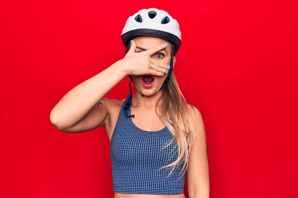 Young beautiful blonde cyclist woman wearing bike security helmet over red background peeking in shock covering face and eyes with hand, looking through fingers afraid
