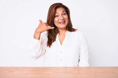 Orta yaşlı esmer, İspanyol iş kadını. Masada oturmuş gülümsüyor. El ve parmaklarıyla telefonla konuşuyor gibi. iletişim kavramları. 