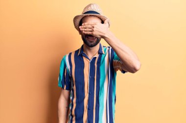 Sakallı, yaz şapkası ve tişörtü giyen, gülümseyen ve elleri yüzünde, şaşırtmak için gözlerini kapatan genç yakışıklı bir adam. kör kavram. 
