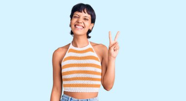 Kısa saçlı, esmer, günlük kıyafetler giyen genç bir kadın. İki numaralı parmaklarıyla işaret ediyor. Kendinden emin ve mutlu gülümsüyor.. 