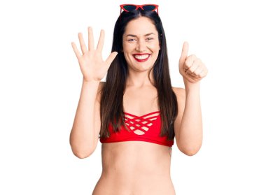 Bikini giyen genç, beyaz bir kadın 6 numaralı parmaklarıyla gösteriş yapıyor ve gülümsüyor. Kendinden emin ve mutlu.. 