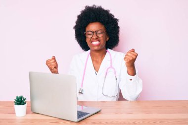 Doktor steteskobu giyen genç Afro-Amerikalı kadın bilgisayar dizüstü bilgisayarı kullanarak çok mutlu ve heyecanlı bir şekilde kollarını kaldırarak, gülümseyerek ve başarı için çığlık atarak kazanan bir jest yapıyor. kutlama konsepti. 