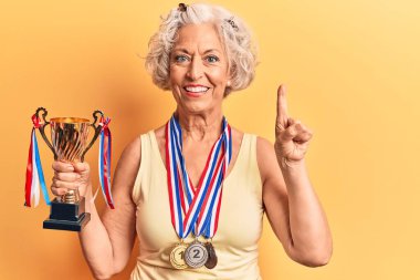 Kıdemli gri saçlı kadın elinde şampiyonluk kupası tutarken bir fikir veya soru işareti ile gülümsüyor. 