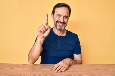 Orta yaşlı İspanyol bir adam gündelik giysiler içinde masada oturuyor ve kendini beğenmiş ve mutlu bir şekilde gülümserken bir numarayı işaret ediyor.. 