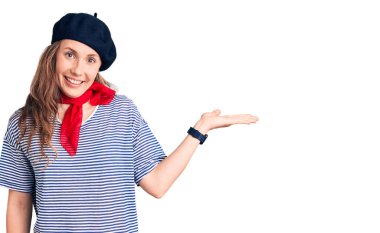 Fransız beresi ve çizgili tişört giyen genç ve güzel sarışın kadın gülümsüyor ve avuç içi kameraya bakıyor.. 