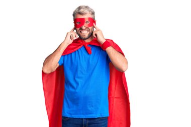 Sarışın, süper kahraman kıyafeti giyen genç bir adam yüksek sesli müzik sesi için kulaklarını parmaklarıyla kapatıyor. sağır kavramı. 