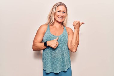 Orta yaşlı beyaz kadın spor giysisi giyiyor. Arka tarafı işaret ediyor. El ve baş parmakları havada, kendine güvenen. 