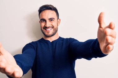 Sakallı, sıradan bir süveter giyen genç yakışıklı beyaz arka planda durup kameraya sarılmak için gülümseyerek bakıyor. Mutluluğu kucaklayan neşeli bir ifade..