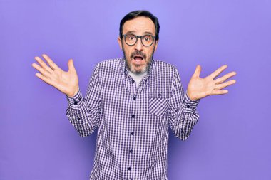 İzole edilmiş beyaz arka plan üzerinde gündelik gömlek ve gözlük takan orta yaşlı yakışıklı bir adam. Kolları açık, hiçbir fikri yok ve şüpheli bir yüzü var..