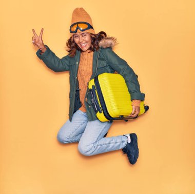 Kışlık giysiler giyen genç, güzel Latin kadın mutlu gülümsüyor. Yüzünde gülümsemeyle kamara çantası tutarak zıplıyor izole edilmiş sarı arka planda zafer işareti yapıyor.