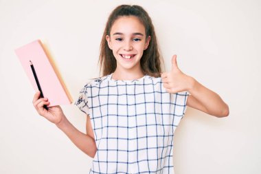 Sevimli İspanyol çocuk elinde kitap ve kalemle gülümsüyor mutlu ve pozitif, başparmağıyla mükemmel ve onay işareti yapıyor. 