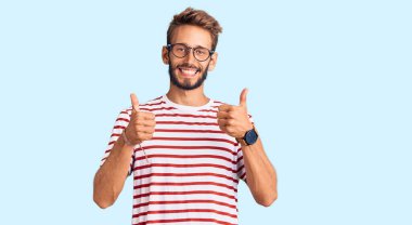 Sakallı, sıradan kıyafetler ve gözlük takan yakışıklı sarışın adam. Başarılı bir el hareketi yapıyor. Gülümsüyor ve mutlu. Neşeli ifade ve kazanan jest. 