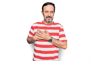 Orta yaşlı, çizgili tişört giyen yakışıklı adam izole edilmiş beyaz arka planda duruyor. Elleri göğsünde gülümsüyor, yüzünde minnet dolu bir jest ile gözleri kapalı. Sağlık kavramı.