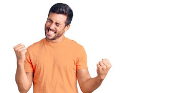 Gündelik kıyafetler giyen genç İspanyol adam çok mutlu ve heyecanlı. Kazanan jestler yapıyor. Kolları havada, gülümsüyor ve başarı için bağırıyor. kutlama konsepti. 