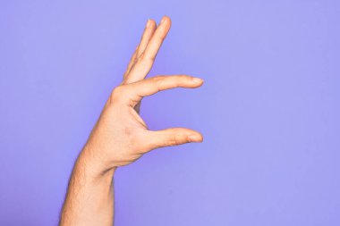 İzole edilmiş mor arka plan üzerinde parmaklarını gösteren beyaz bir adamın eli. Görünmez bir şeyi alıyor, parmakları boşluğu gösteren bir nesneyi tutuyor.