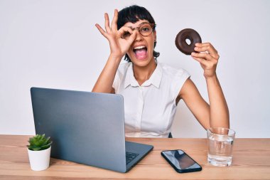 Ofiste çalışan güzel bir esmer kadın çikolatalı donut yiyor gülümsüyor ve mutlu mesut gülümsüyor. 