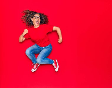 Orta yaşlı, güzel İspanyol bir kadın günlük kıyafetler ve mutlu bir gülümseme takınıyor. İzole edilmiş kırmızı arka planda yüzünde gülümsemeyle zıplıyor