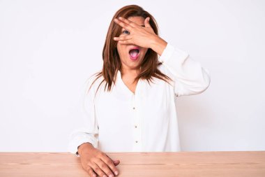 Orta yaşlı esmer, Latin kökenli, sıradan beyaz gömlekli bir kadın masanın üzerinde oturuyor. Şok olmuş bir şekilde yüzünü ve gözlerini kapatıyor. Utanmış bir ifadeyle parmaklarının arasından bakıyor.. 
