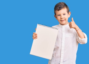 Sevimli sarışın çocuk elinde defterle mutlu ve pozitif gülümsüyor, başparmağıyla mükemmel ve onay işareti yapıyor. 