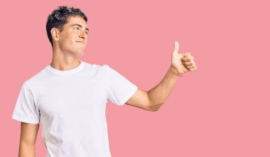 Genç ve yakışıklı bir adam günlük beyaz tişört giyiyor. Gururlu görünüyor, gülümsüyor. Kenarda baş parmak hareketi yapıyor. 