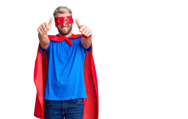 Sarışın, süper kahraman kıyafeti giymiş genç bir adam elleriyle olumlu bir jest yapmayı onaylıyor, gülümsüyor ve başarı için mutlu. kazanan jest. 
