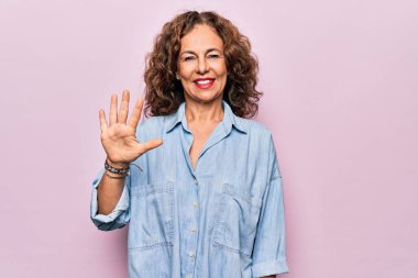 Orta yaşlı güzel bir kadın pembe arka planda dikilen sıradan bir kot gömlek giyer ve parmaklarıyla 5 numarayı gösterirken kendinden emin ve mutlu bir şekilde gülümser..