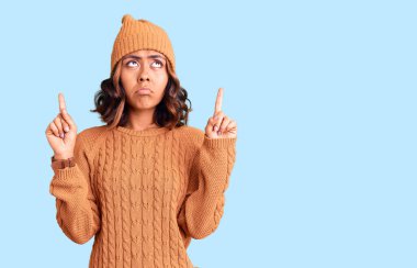 Genç, güzel, melez, yün kazak giyen ve kışlık şapkalı bir kadın üzgün ve üzgün görünüyor, parmaklarıyla yön gösteriyor, mutsuz ve depresif.. 