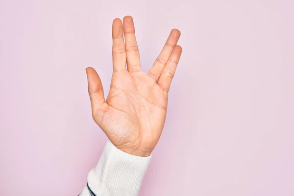 Hand of caucasian young man showing fingers over isolated pink background greeting doing Vulcan salute, showing hand palm and fingers, freak culture