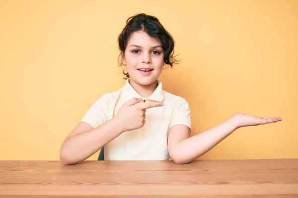 テーブルの上に座っているカジュアルな服を着てかわいいヒスパニック系の子供は 手で提示し 指で指している間 カメラに驚いて笑顔 — ストック写真
