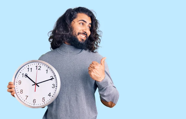 Młody Arabski Człowiek Trzymając Duży Zegar Wskazując Kciuk Górę Bok — Zdjęcie stockowe