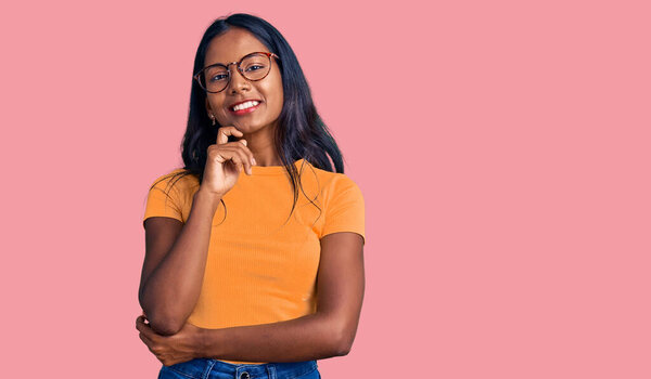 Young indian girl wearing casual clothes and glasses looking confident at the camera with smile with crossed arms and hand raised on chin. thinking positive. 
