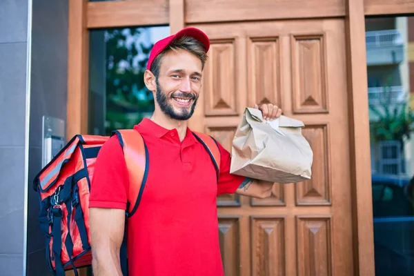 赤い制服と配送バックパックを身に着けている白人の配達人笑顔幸せな屋外保持紙バッグ — ストック写真